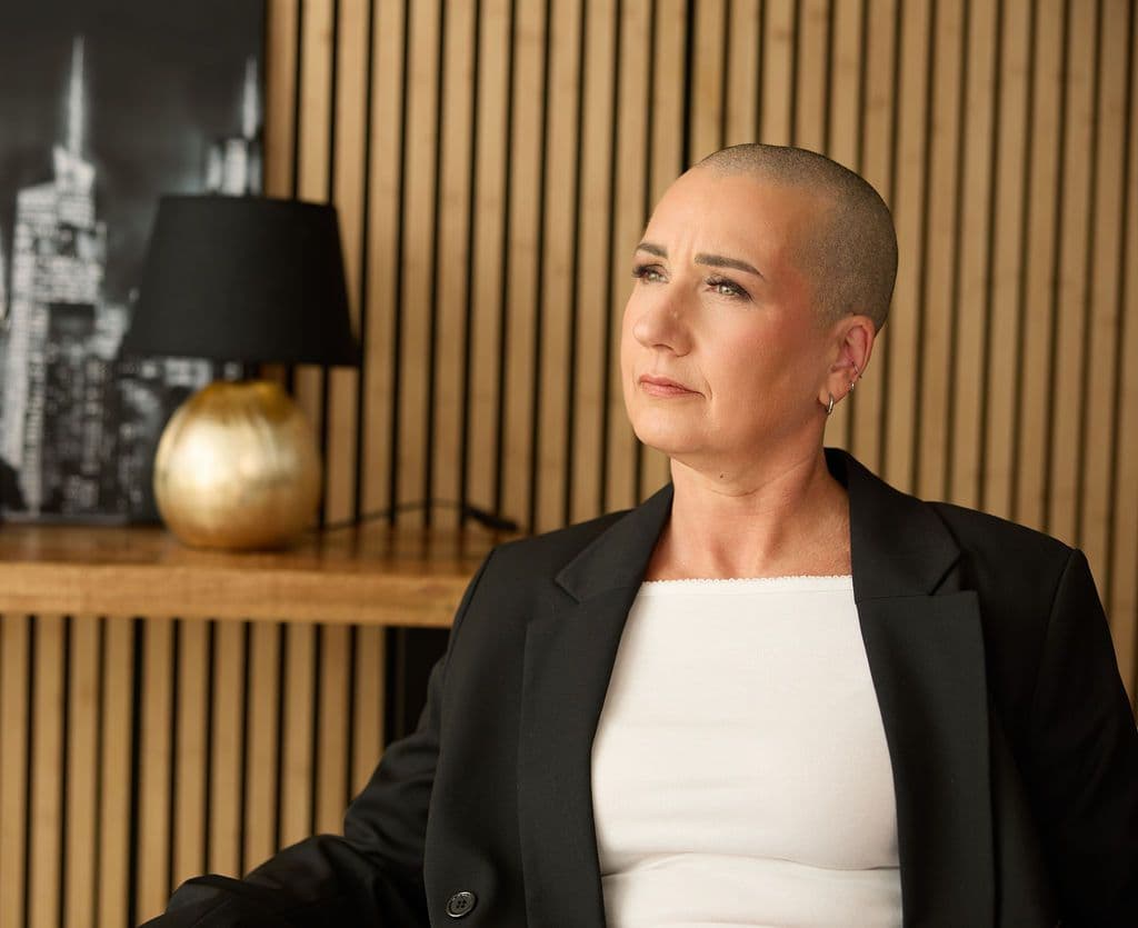 Jana Schulze im schwarzen Blazer vor einer Holzwand, den Blick nach links gerichtet.