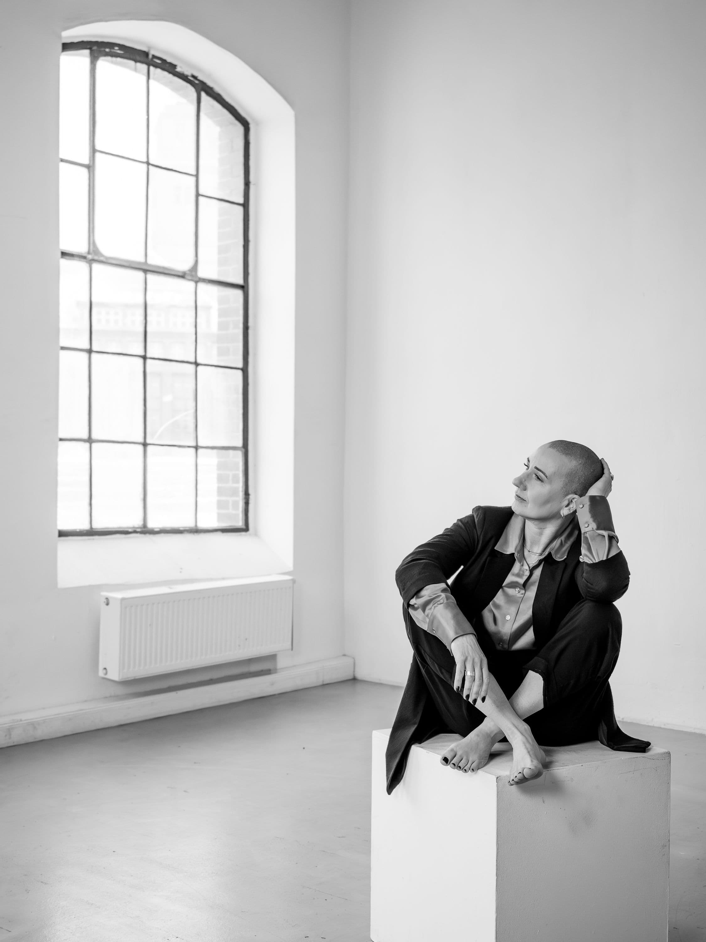 Jana Schulze sitzt barfuß auf einem weißen Podest und blickt zu einem großen Industriefenster.